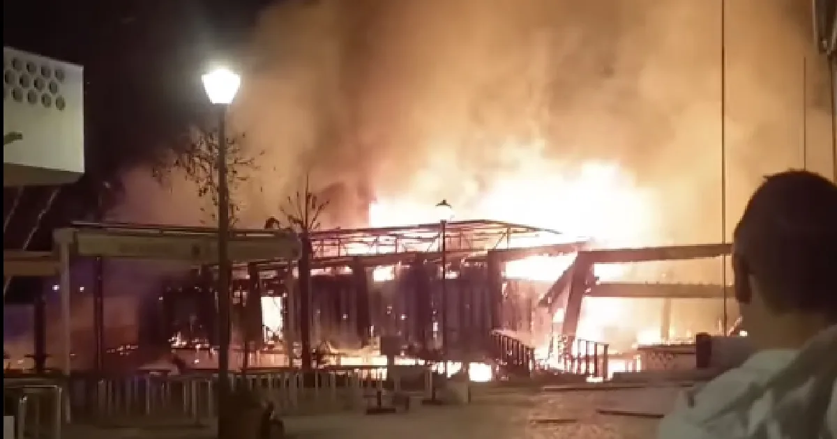 Incêndio intenso consome edifício à noite, observador em primeiro plano.